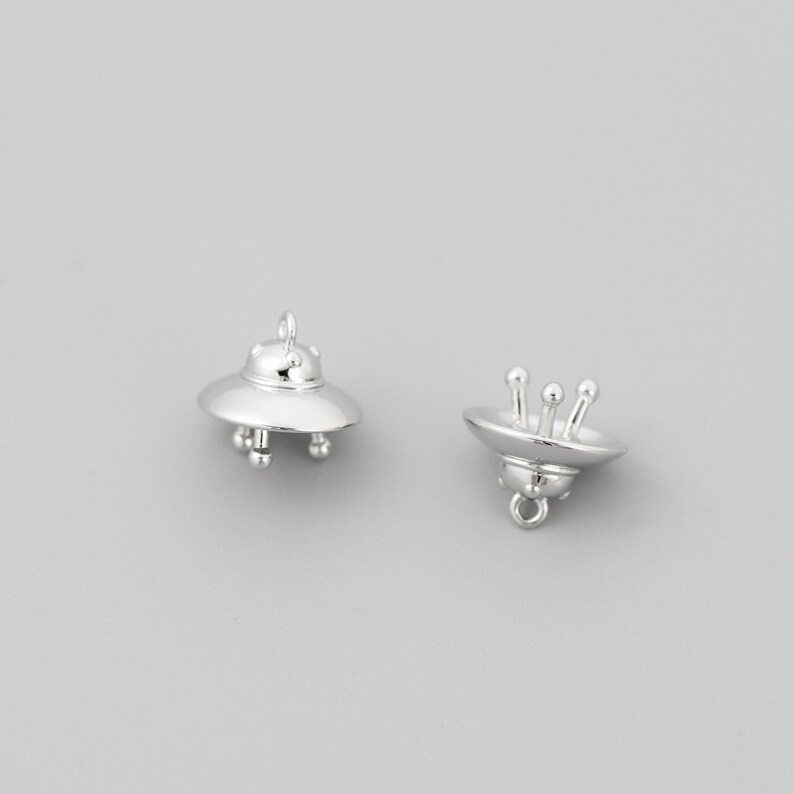 Op de afbeelding: Twee zilveren UFO-bedels. Elke bedel heeft een gedetailleerd UFO-ontwerp met een afgeronde bovenkant en een platte onderkant. E&eacute;n bedel heeft een lus aan de bovenkant om aan een ketting of armband te bevestigen. De andere bedel heeft een lus aan de onderkant.