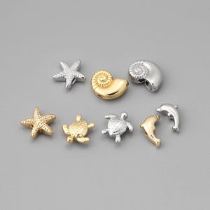 May include: A collection of small, metallic charms in various sea-themed shapes. The charms include starfish, seashells, turtles, and dolphins, in gold and silver tones. These decorative charms are ideal for jewelry making or craft projects.