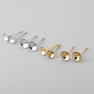 Peut inclure: Une collection de boucles d'oreilles à clous argentées et dorées. Les boucles d'oreilles ont une tige et une petite coupelle ronde et peu profonde. Les boucles d'oreilles sont présentées sur une surface gris clair.