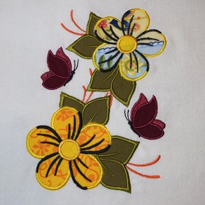 Peut inclure: Broderie représentant deux fleurs jaunes avec des contours noirs et un cercle jaune au centre, entourées de feuilles vertes et de deux papillons bordeaux.