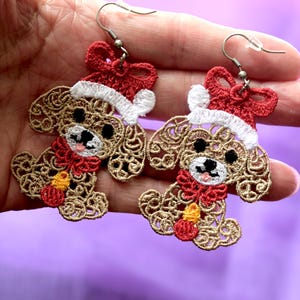May include: A pair of dog-shaped earrings with a festive theme. The earrings are made of tan-coloured lace, each depicting a dog wearing a red Santa hat and scarf. They have black eyes, a pink mouth, and a small yellow candle.