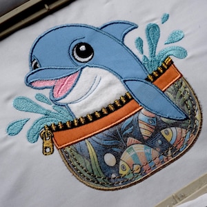 Peut inclure: Écusson brodé représentant un dauphin bleu souriant sortant d'une poche. La poche, avec une fermeture éclair, contient des poissons colorés et des plantes aquatiques. Le dauphin est entouré d'éclaboussures d'eau bleues.