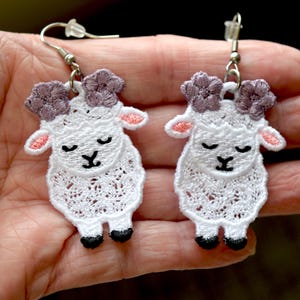 May include: A pair of white lace sheep earrings with lavender horns and pink inner ears. The sheep have black eyes and feet. The earrings are held by silver hooks.