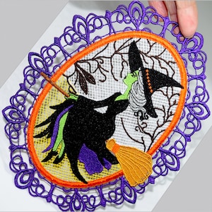 Könnte beinhalten: Gestickte ovale Halloween-Tischdecke mit einer Hexe auf einem Besen. Die Hexe ist in Schwarz, Grün und Weiß dargestellt, mit einem gelben Besen. Das Design wird von einem orangefarbenen Rand und einem lila Spitzenrand umrahmt.