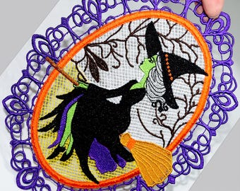 May include: Embroidered Halloween-themed oval doily featuring a witch on a broom. The witch is depicted in black, green, and white, with a yellow broom. The design is framed by an orange border and a purple lace edge.