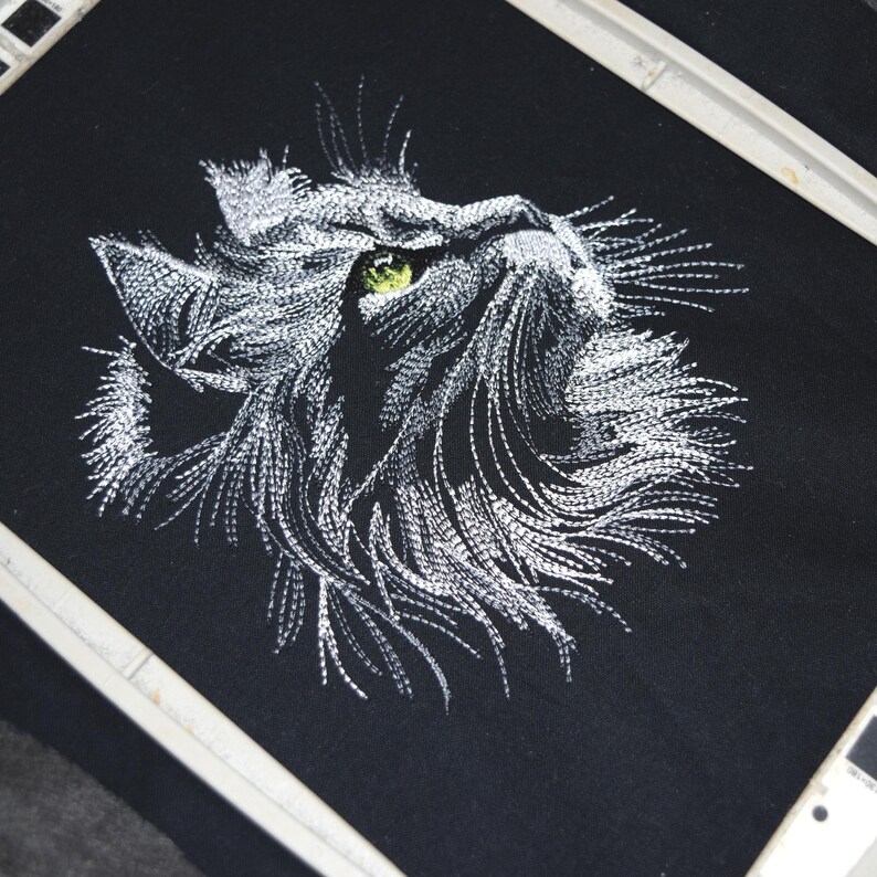 May include: Embroidered cat face on black fabric. The cat's face is rendered in white thread, with a detailed depiction of fur and a single green eye. The embroidery is framed by a white hoop, suggesting a craft project.