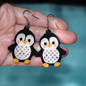 Peut inclure: Une paire de boucles d'oreilles en forme de pingouin. Les boucles d'oreilles présentent des pingouins brodés noirs et blancs avec des pieds et des becs orange. Les pingouins se tiennent la main et ont des joues roses. Les boucles sont suspendues à des crochets argentés.