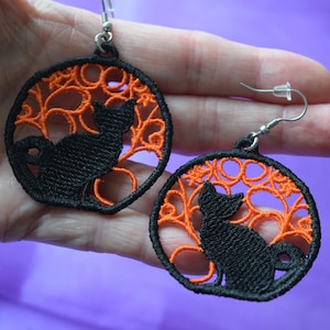 May include: Black cat earrings with orange filigree design. The earrings feature a black cat silhouette within a circular frame of orange embroidery. The earrings have silver-colored hooks.