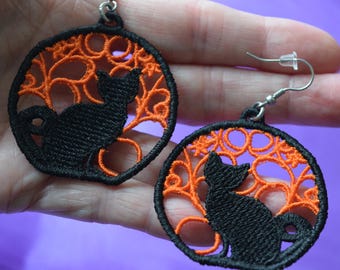 Puede incluir: Pendientes de gato negro bordados con un diseño de filigrana naranja. Los pendientes presentan una silueta de gato negro dentro de un marco circular de bordado naranja. Los pendientes tienen ganchos plateados.