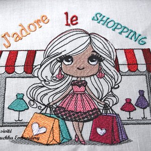 Op de afbeelding: Geborduurd ontwerp met een cartoonmeisje met lang wit haar, met boodschappentassen. De afbeelding bevat de tekst "J'adore le SHOPPING" en een winkelmarkies. Het meisje draagt een roze jurk en oorbellen.