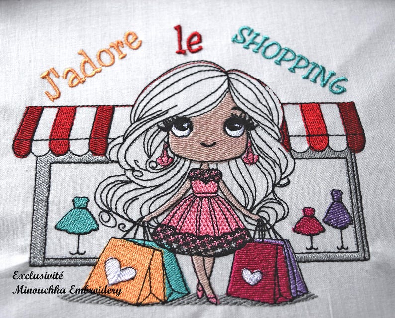 Op de afbeelding: Geborduurd ontwerp met een cartoonmeisje met lang wit haar, met boodschappentassen. De afbeelding bevat de tekst "J'adore le SHOPPING" en een winkelmarkies. Het meisje draagt een roze jurk en oorbellen.