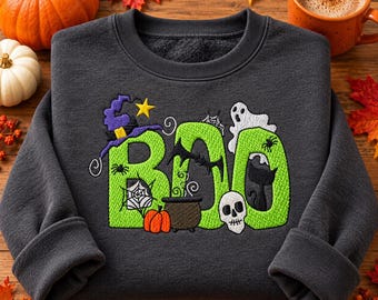 May include: A dark gray crewneck sweatshirt featuring a large embroidered design of the word BOO in bright green letters. The design includes a purple witch hat, a yellow star, a white ghost, a black cat, a skull, and a small cauldron.