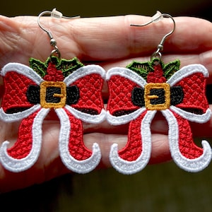 May include: A pair of Christmas earrings. Each earring is a red bow with white trim, a black belt, and a gold buckle. Green holly leaves and silver earring hooks complete the festive design.