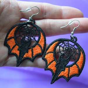 Op de afbeelding: Een paar Halloween-oorbellen. Elke oorbel heeft een zwart vleermuissilhouet in een oranje vleermuisvleugelontwerp, omringd door een zwart spinnenweb. De oorbellen hebben zilverkleurige oorhaken.