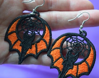 Puede incluir: Un par de pendientes con temática de Halloween. Cada pendiente presenta una silueta de murciélago negro dentro de un diseño de ala de murciélago naranja, rodeada por una telaraña negra. Los pendientes tienen ganchos para las orejas plateados.