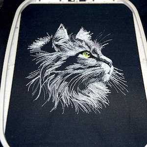 May include: An embroidered cat profile in white thread on black fabric. The cat's eye is a bright green, and the embroidery is detailed, showing the texture of the fur. The fabric is stretched in an embroidery hoop.