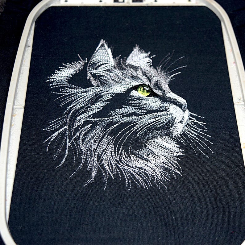May include: An embroidered cat profile in white thread on black fabric. The cat's eye is a bright green, and the embroidery is detailed, showing the texture of the fur. The fabric is stretched in an embroidery hoop.