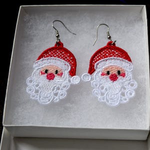 Pode incluir: Um par de brincos pendurados com tema de Papai Noel. Cada brinco apresenta um chapéu vermelho com acabamento branco, um rosto rosa e uma barba branca. Os brincos são feitos de tecido bordado e têm ganchos prateados.