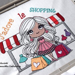 Op de afbeelding: Geborduurd ontwerp met een cartoonmeisje met lang wit haar, met boodschappentassen. Het ontwerp bevat de woorden "J'adore le SHOPPING" en "Exclusivit&eacute; Minouchka Embroidery". Het meisje draagt een roze jurk en staat voor een winkel.