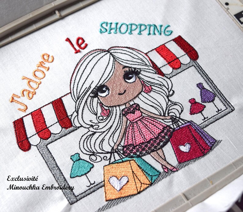Op de afbeelding: Geborduurd ontwerp met een cartoonmeisje met lang wit haar, met boodschappentassen. Het ontwerp bevat de woorden "J'adore le SHOPPING" en "Exclusivit&eacute; Minouchka Embroidery". Het meisje draagt een roze jurk en staat voor een winkel.