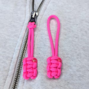 May include: Two pink zipper pulls with a knotted design. The zipper pulls are attached to a zipper on a grey hoodie.