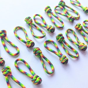 May include: A collection of colorful zipper pulls. Each pull features a loop of rainbow-colored cord attached to a knotted end and a small, dark-colored stopper. The pulls are arranged on a white surface.