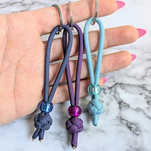 May include: Three keychains in shades of blue and purple. Each keychain features a knotted cord loop, a decorative bead, and a metal ring. The keychains are in varying shades of blue and purple, with a light blue, a dark blue, and a purple.