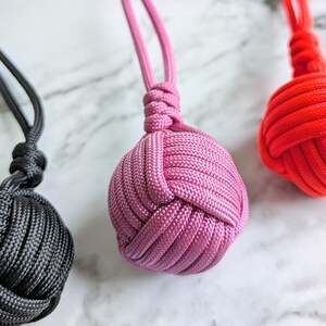 Peut inclure: Trois porte-clés en paracorde faits à la main avec des nœuds de différentes couleurs. Les porte-clés sont noir, rose et orange.