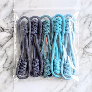 May include: A set of six blue and gray paracord zipper pulls. The zipper pulls are made with a knotting technique and have a gradient color pattern.