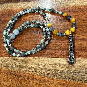 May include: A beaded necklace featuring a variety of stone beads in shades of gray, turquoise, yellow, and white. The necklace has a silver-colored pendant with a textured design. The beads are strung together to create a layered effect.
