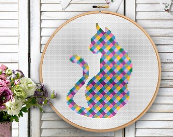 May include: A cross-stitch cat design in a wooden hoop. The cat is made up of a colourful geometric pattern in pink, yellow, blue, and green. The background features a white shutter and floral bunting.