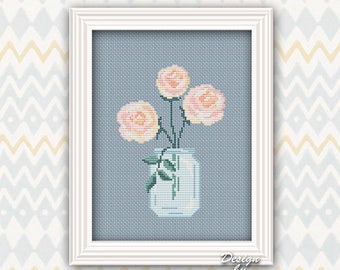 May include: Cross-stitch art of three peach-colored roses in a white jar, set against a light blue background. The artwork is framed in a white rectangular frame. The design is by Modern Cross Stitch Art.