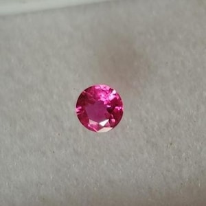 May include: A round, faceted, hot pink gemstone, possibly a ruby or pink sapphire, set against a white background within a white plastic container. The gemstone is the focal point, showcasing its brilliant cut and vibrant colour.