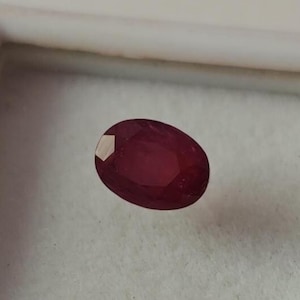 May include: A faceted, oval-shaped ruby gemstone. The ruby is a deep, rich red color and is set against a white background within a white container. The gemstone has a polished surface with visible facets.