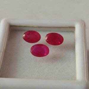 May include: Three oval-cut, vibrant red gemstones are displayed within a white, square-shaped container. The gems have a polished, reflective surface, showcasing their rich color and clarity. The arrangement highlights the gemstones' individual beauty.