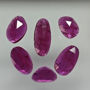 Puede incluir: Seis piedras preciosas ovaladas de color rosa con bordes facetados. Las piedras preciosas están dispuestas en semicírculo sobre una superficie blanca.