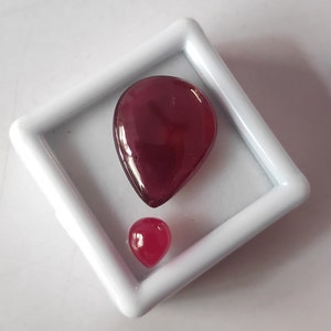 May include: Two red gemstone cabochons in a white square display box. The larger cabochon is teardrop shaped and the smaller cabochon is round.