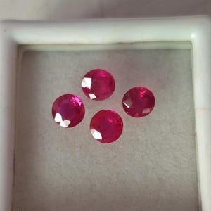 Könnte beinhalten: Vier runde, facettierte, leuchtend pink-rote Edelsteine, die in einer quadratischen Anordnung in einer weißen Schachtel präsentiert werden. Jeder Stein hat einen Brillantschliff mit zahlreichen Facetten, die das Licht reflektieren.