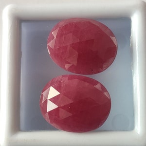 Pode incluir: Duas pedras preciosas ovais vermelhas com corte facetado. As pedras preciosas são exibidas em uma caixa branca.