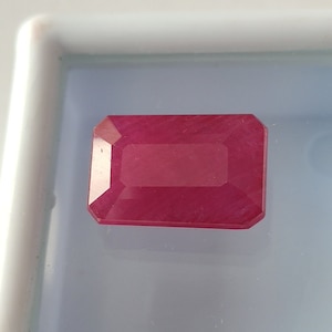May include: A deep red gemstone with a rectangular emerald cut. The gemstone is faceted and has a polished finish.