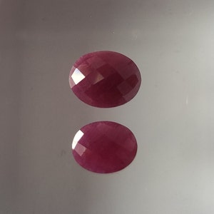 May include: Two round, faceted, ruby red gemstones. The larger gemstone is on top of the smaller gemstone.