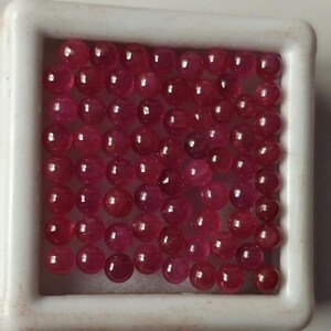 May include: A collection of small, round, polished ruby gemstones in a white square tray. The rubies are a deep red color with a glossy finish, and they are densely packed within the tray. The gemstones are likely for jewelry making or collecting.