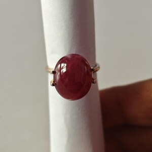 Op de afbeelding: Een gouden ring met een grote, gladde, ovaalvormige dieprode cabochon edelsteen. De steen wordt stevig op zijn plaats gehouden door vier kleine pootjes op een dunne, gepolijste gouden band, wat een eenvoudig en elegant sieraad vormt.