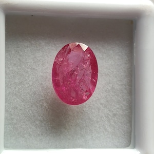 Puede incluir: Una piedra preciosa ovalada de color rosa con un corte facetado. La piedra preciosa tiene una superficie rugosa y texturizada.