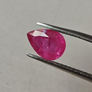 Może przedstawiać: Szlifowany rubin w kształcie gruszki o intensywnym czerwonym kolorze. Kamień jest trzymany w pęsecie.