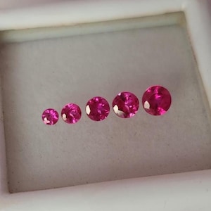 May include: Five round, faceted, vibrant pink gemstones arranged in a horizontal row from smallest to largest. Each stone features a brilliant cut with multiple facets that reflect light, displayed inside a white square storage container.