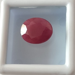 Pode incluir: Uma gema oval de cor vermelha profunda, provavelmente um rubi, é exibida em uma caixa quadrada branca.
