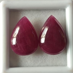 May include: Two teardrop-shaped, deep pink gemstones in a white box. The gemstones are smooth and shiny.