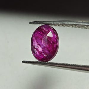 May include: A vibrant pink oval-shaped gemstone with a faceted cut. The gemstone is held by a pair of tweezers.