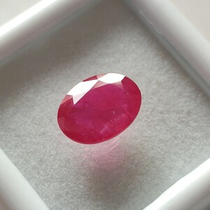 May include: A vibrant red oval-shaped gemstone with a faceted cut. The gemstone is displayed in a white box.